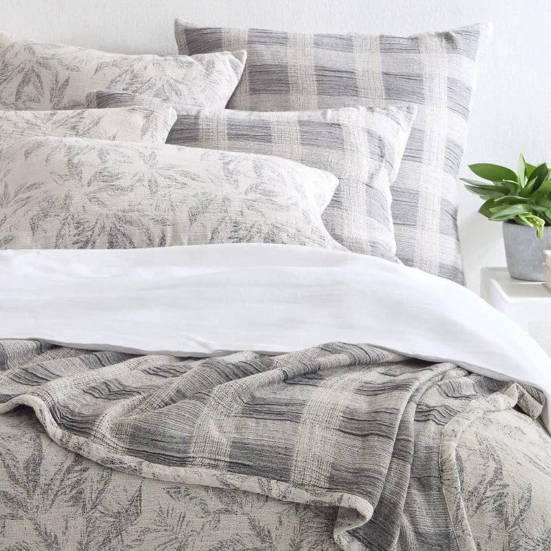 A bed with a gray floral matelassé coverlet, matching pillows, and a plaid coverlet underneath. The room has a light interior with a plant on the nightstand and a window in the background.